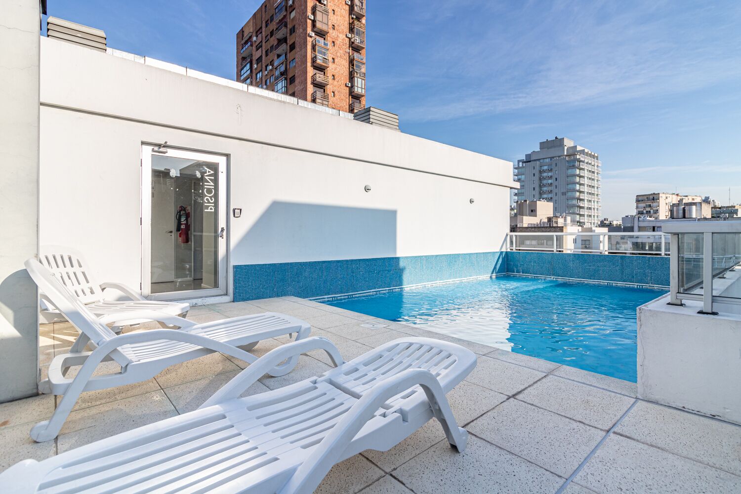 A rooftop pool area with white lounge chairs, a glass door entrance, and a blue-tiled pool.