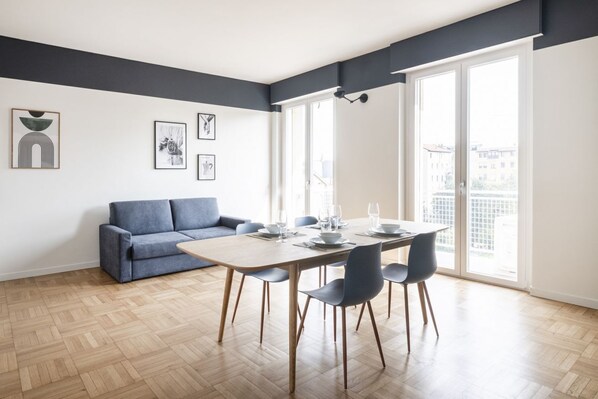 Una sala de estar moderna con una mesa de comedor dispuesta para cuatro personas, un sofá azul y ventanas grandes con vista a la ciudad.