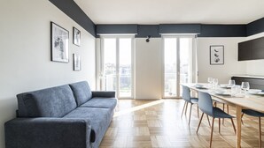 Un salon moderne avec un canapé gris, un coin repas doté d’une table et de chaises en bois, et de grandes fenêtres offrant une vue sur la ville.