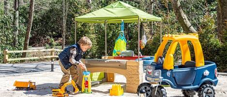 A child playing with toy vehicles and a sandbox in an outdoor play area.
