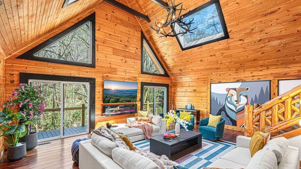 A spacious wooden cabin with a high ceiling, large windows, a cozy living area with a sofa and coffee table, and a staircase leading to an upper level.
