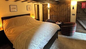 Une chambre avec un grand lit, des poutres en bois, un fauteuil à carreaux et un escalier menant à une mezzanine.