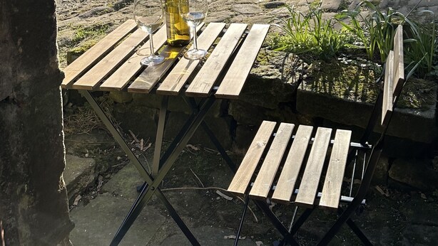 Une table en bois, avec une bouteille de vin et des verres, placée devant un mur de pierre, avec vue sur un bâtiment en pierre et des arbres.