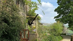 Un étroit sentier de gravier mène à un portail en bois rustique, entouré d’un feuillage dense et d’un bâtiment en pierre.
