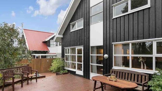 Una casa moderna con fachada negra, detalles blancos y techo rojo. Cuenta con una terraza de madera con bancos y una mesa, rodeada de vegetación.
