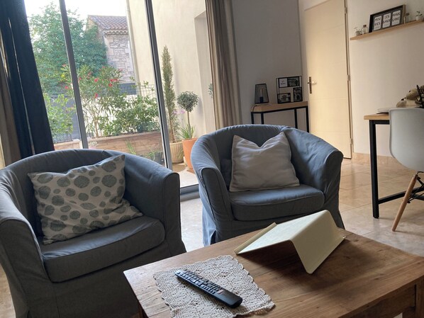 A modern living room with two grey armchairs, a wooden coffee table with a remote control and a book, and a view of a garden through large windows.