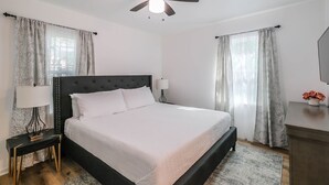 Ein ordentlich eingerichtetes Schlafzimmer mit Bett, Nachttischlampen, Deckenventilator und Vorhängen.