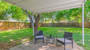 Ein überdachter Patio-Bereich mit zwei Korbstühlen und einem kleinen Tisch, umgeben von einem Holzzaun und Bäumen.