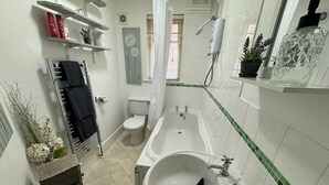 A bathroom with a toilet, bathtub, and sink. There are shelves with toiletries and a shower area. - Cosy 3-bed Apartment in Hythe High Street (Hythe)