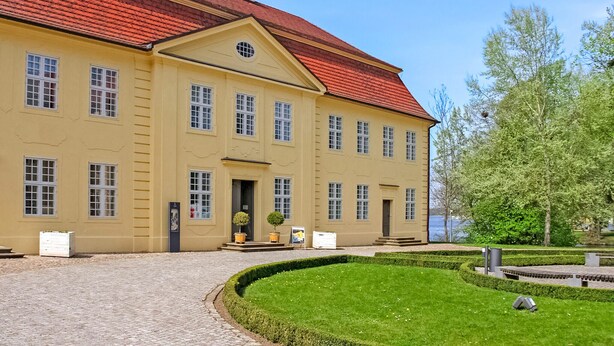 A large, yellow building with a red roof, multiple windows, and a well-maintained garden with a circular hedge and a small tree. - 2 bedroom lovely apartment in Mirow (Mirow)