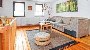 A living room with a grey sofa, wooden flooring, a round wooden table, a woven ottoman, and a forest-themed wall art. - 2 bedroom lovely apartment in Mirow (Mirow)