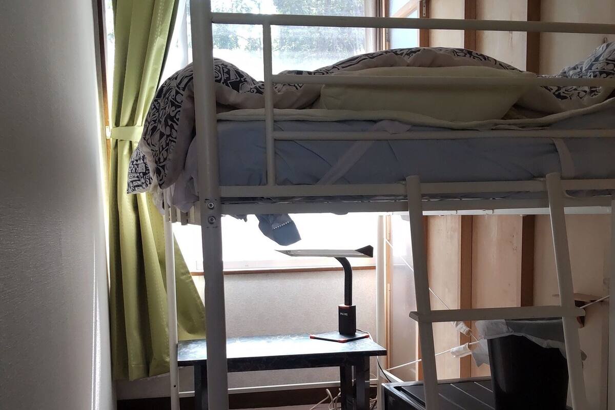 Quarto com beliche, escrivaninha e cadeira, janela com cortinas e vista para árvores.