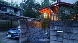 Una entrada con portón, un automóvil estacionado adentro, un edificio con techo rojo y un letrero con caracteres japoneses.