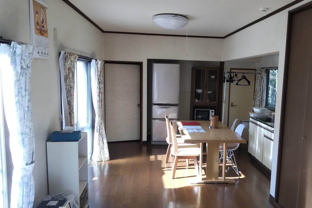 Quarto com mesa de jantar de madeira, cadeiras, frigorífico, micro-ondas e armário com pratos.