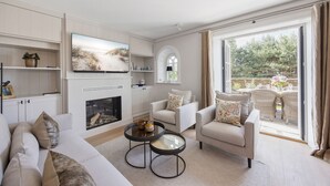 A modern living room with a fireplace, comfortable seating, a flat-screen TV, and a large window offering a view of the outdoors. - Neubau. Wellness. Reet (List auf Sylt)