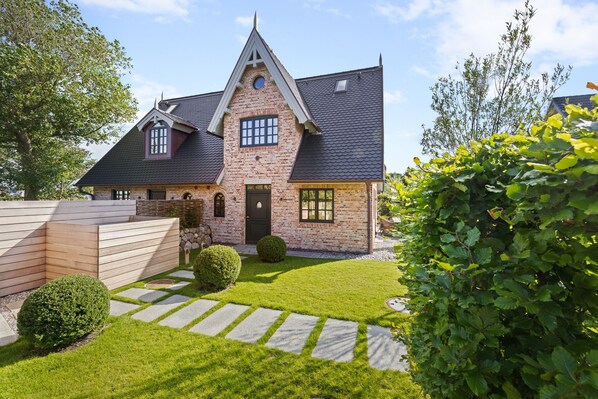 A two-story house with a steeply pitched roof, brick exterior, and a well-maintained lawn with stone pathways. - Neubau. Familie. Bäderstil (List auf Sylt)