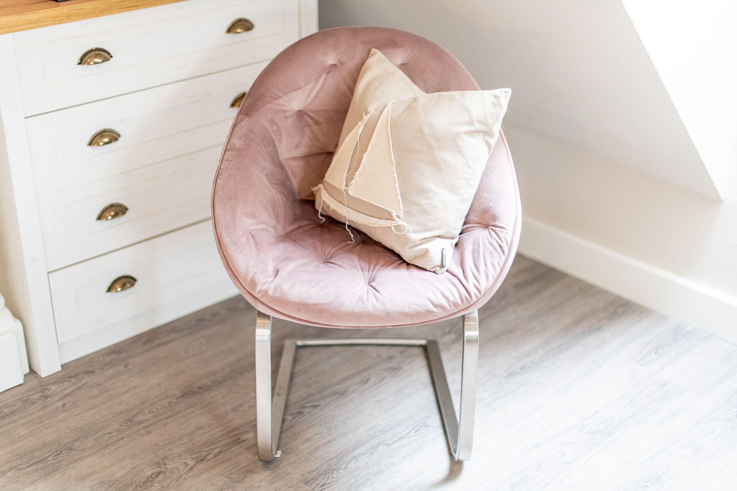 Een roze bekleden stoel met een beige kussen, geplaatst naast een witte ladekast met messing handgrepen.