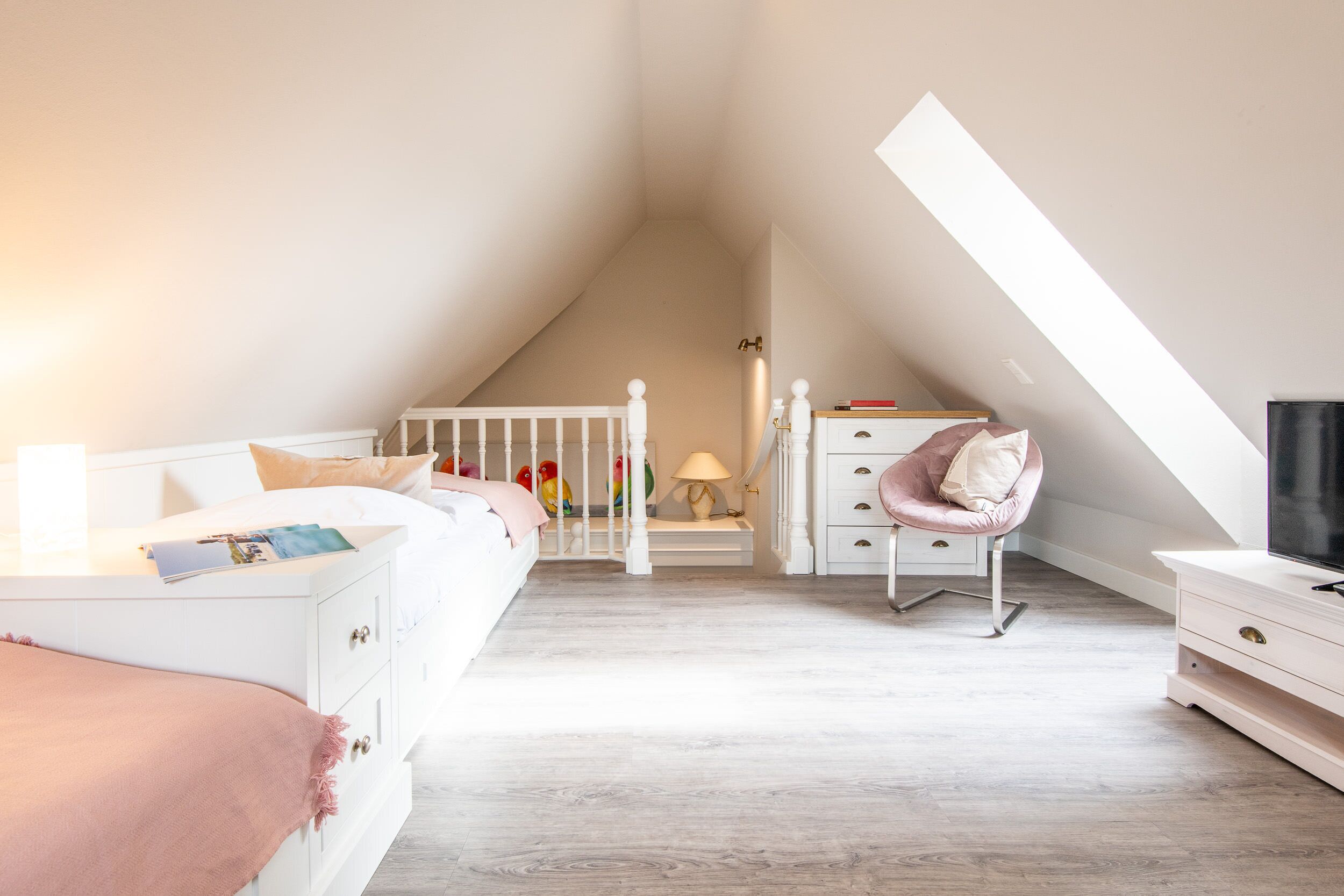 Une chambre avec un plafond en pente, un lit recouvert d’une couverture rose, une chaise, une commode et un téléviseur.