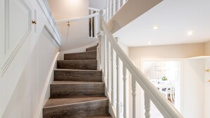 A staircase with wooden steps and white railings, leading to a room with a white door and a dining area visible in the background. - Gemütlich. Ruhig. Garten (Sylt)