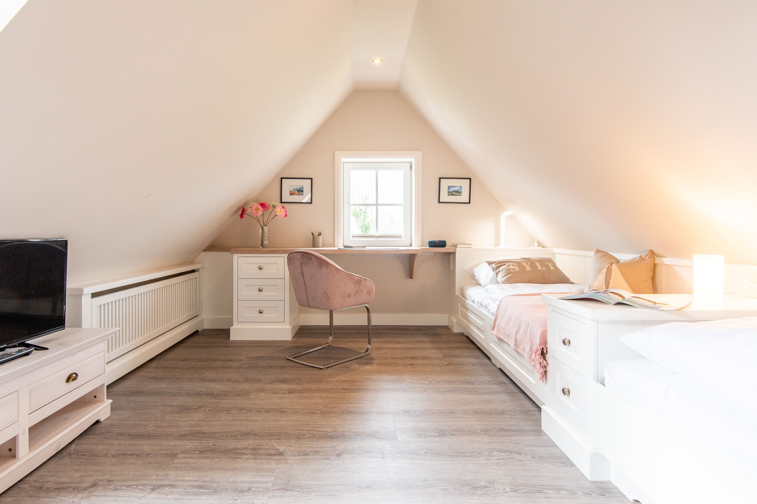 A bedroom with a sloped ceiling, a white dresser, a pink chair, a bed with pillows, a window, and a radiator.