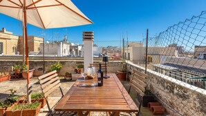 Una mesa de madera con una botella de vino y copas, rodeada de plantas en macetas, bajo un paraguas beige, con vistas a edificios a través de una valla de malla.
