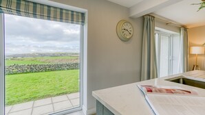 Una habitación con un ventanal amplio que ofrece vistas a un campo de césped y un muro de piedra, un reloj en la pared y una mesa con un libro.