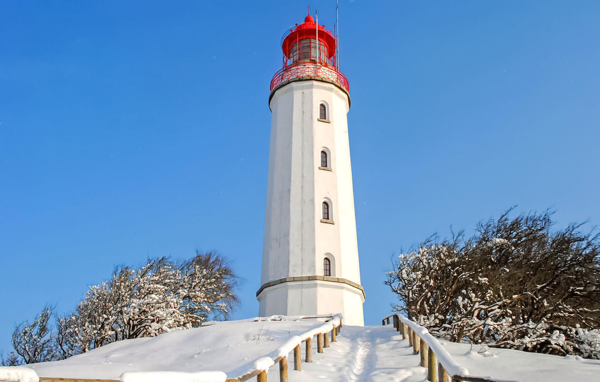 Un alto faro con la cima rossa, circondato da alberi innevati e un cielo azzurro limpido.