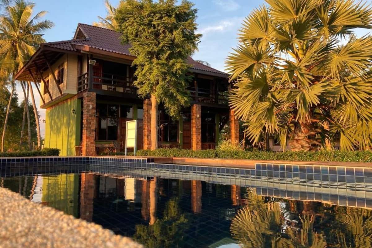 A two-story house with a tiled roof, surrounded by palm trees and a swimming pool.
