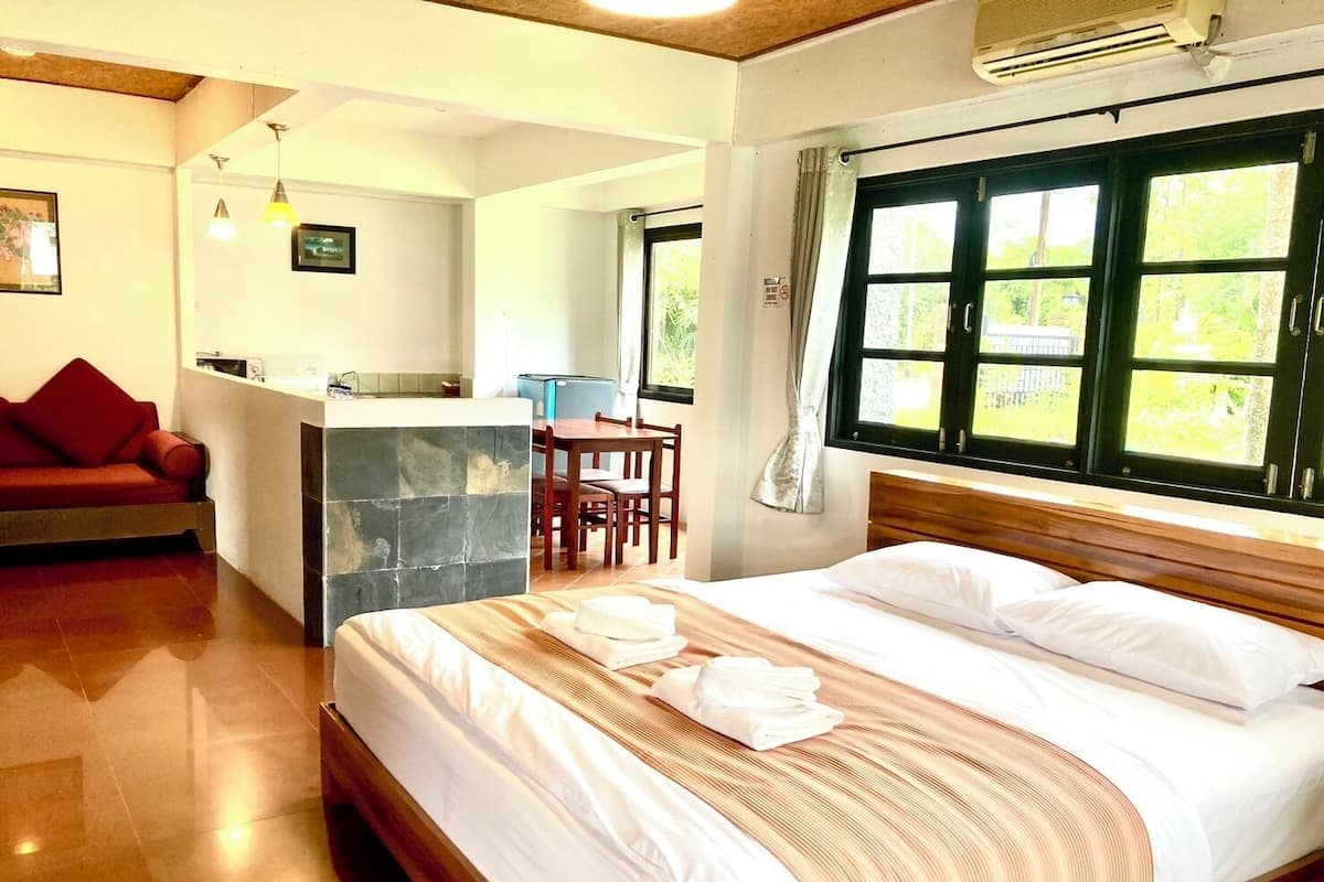 A hotel room with a bed, a small kitchenette, a dining area, and a window with a view of greenery.