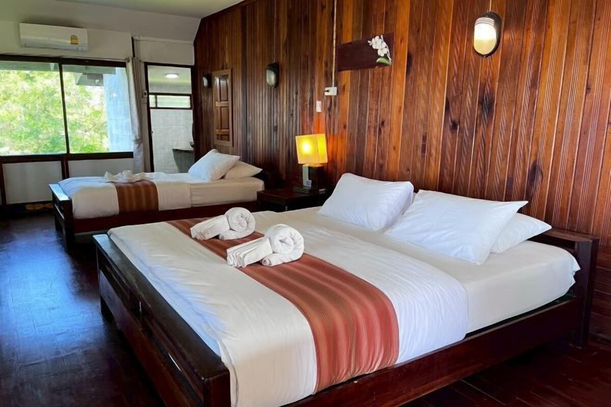 A hotel room with two beds, wooden paneling, a nightstand with a lamp, and a window with curtains.