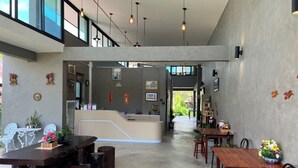 A spacious, modern interior with a central reception desk, wooden tables, and chairs, surrounded by artwork and potted plants. - The Chevalley beach resort  (Thung Wa)