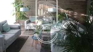 Ein modernes Wohnzimmer mit einem grauen Sofa, einem Couchtisch und einer Lampe. Im Hintergrund sind Pflanzen und eine Holzwand zu sehen.