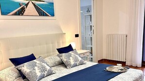 Ein geräumiges Schlafzimmer mit einem großen Bett, Blick auf Strand und Steg durch ein Fenster, ein Badezimmer mit Glastür und eine weiße Klimaanlage.