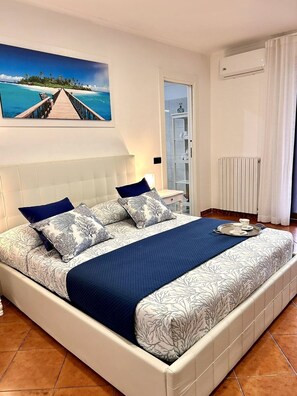 Un'ampia camera da letto con un grande letto, vista su una spiaggia e un molo attraverso una finestra, bagno con porta in vetro e condizionatore d'aria bianco.