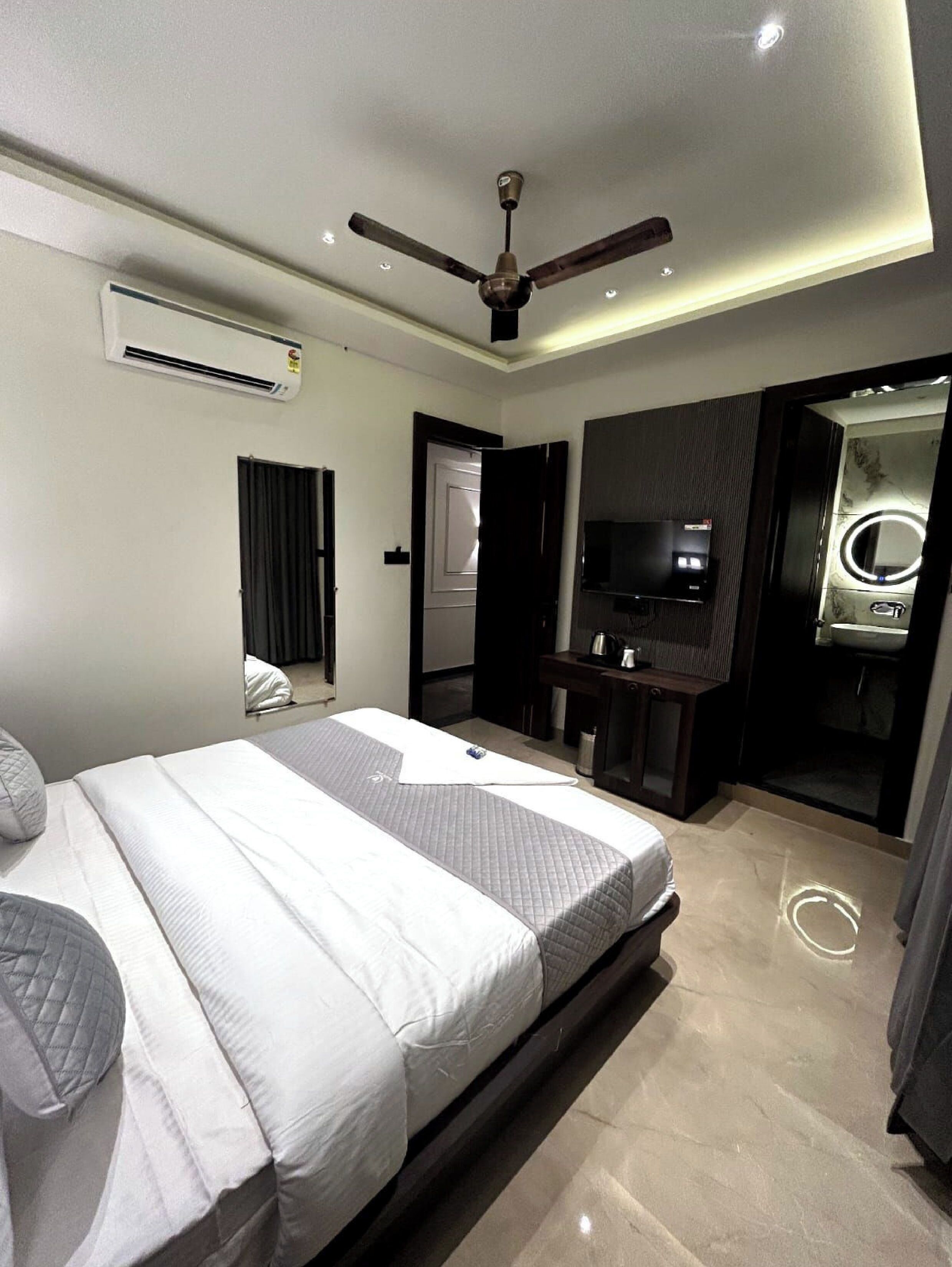 A modern hotel room with a bed, ceiling fan, air conditioning unit, TV, and a bathroom with a mirror and sink.