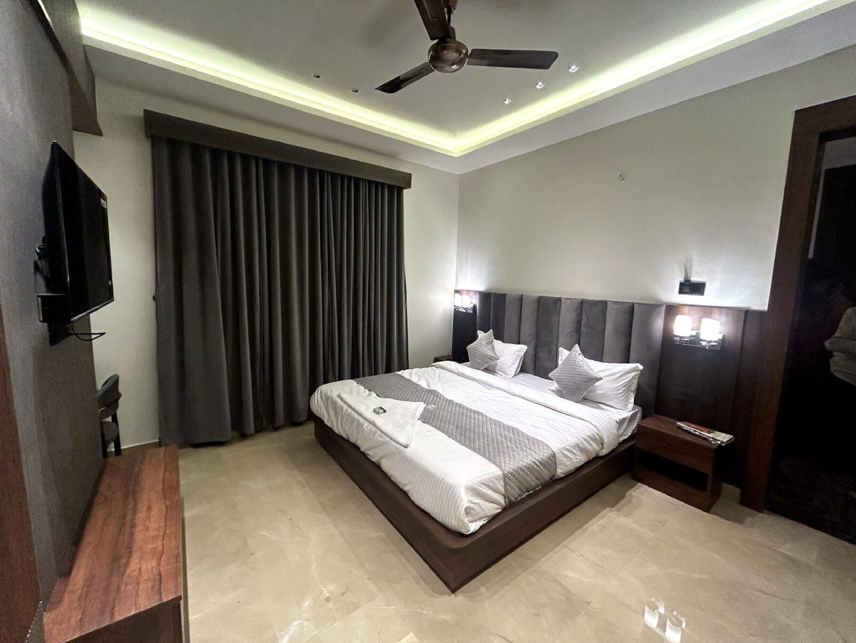 A modern hotel room with a large bed, a ceiling fan, a TV, and a nightstand with a lamp.
