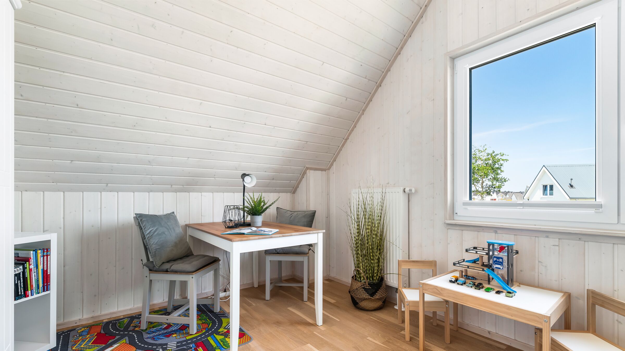 A cozy attic room with a sloped ceiling, a wooden table, two chairs, a bookshelf, a window with a view of houses, and a toy train set.