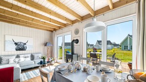 Ein gemütlicher Raum mit Holzboden, einem grauen Sofa, einem Esstisch mit weißem Geschirr und Gläsern sowie einem großen Fenster mit Blick auf Häuser.