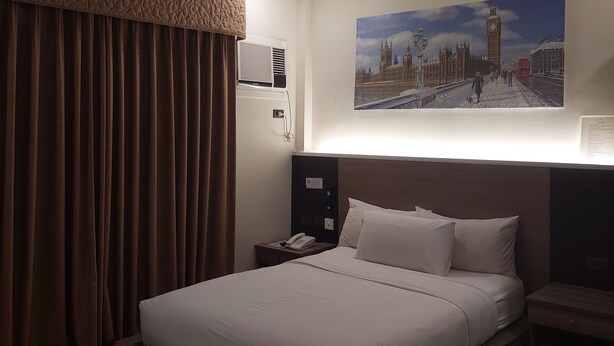 Een hotelkamer met een groot bed, een nachtkastje met telefoon, een schilderij van een stadsgezicht en een aan het plafond bevestigde airconditioning.