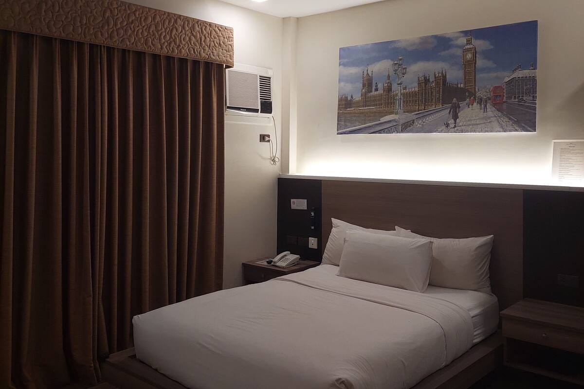 A hotel room with a large bed, a nightstand with a phone, a painting of a cityscape, and a ceiling-mounted air conditioning unit.