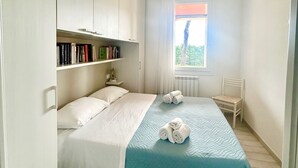 A small, well-lit bedroom with a bed, a window with curtains, a chair, and a bookshelf. - Garden Retreat by the sea on Albarella (Rosolina)