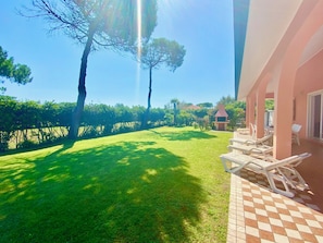 A patio with lounge chairs, a tiled floor, and a view of a grassy area with trees and a building in the background. - Garden Retreat by the sea on Albarella (Rosolina)