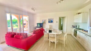A bright living room with a red sofa, a dining table with chairs, a TV, and a kitchen with cabinets and appliances. - Garden Retreat by the sea on Albarella (Rosolina)