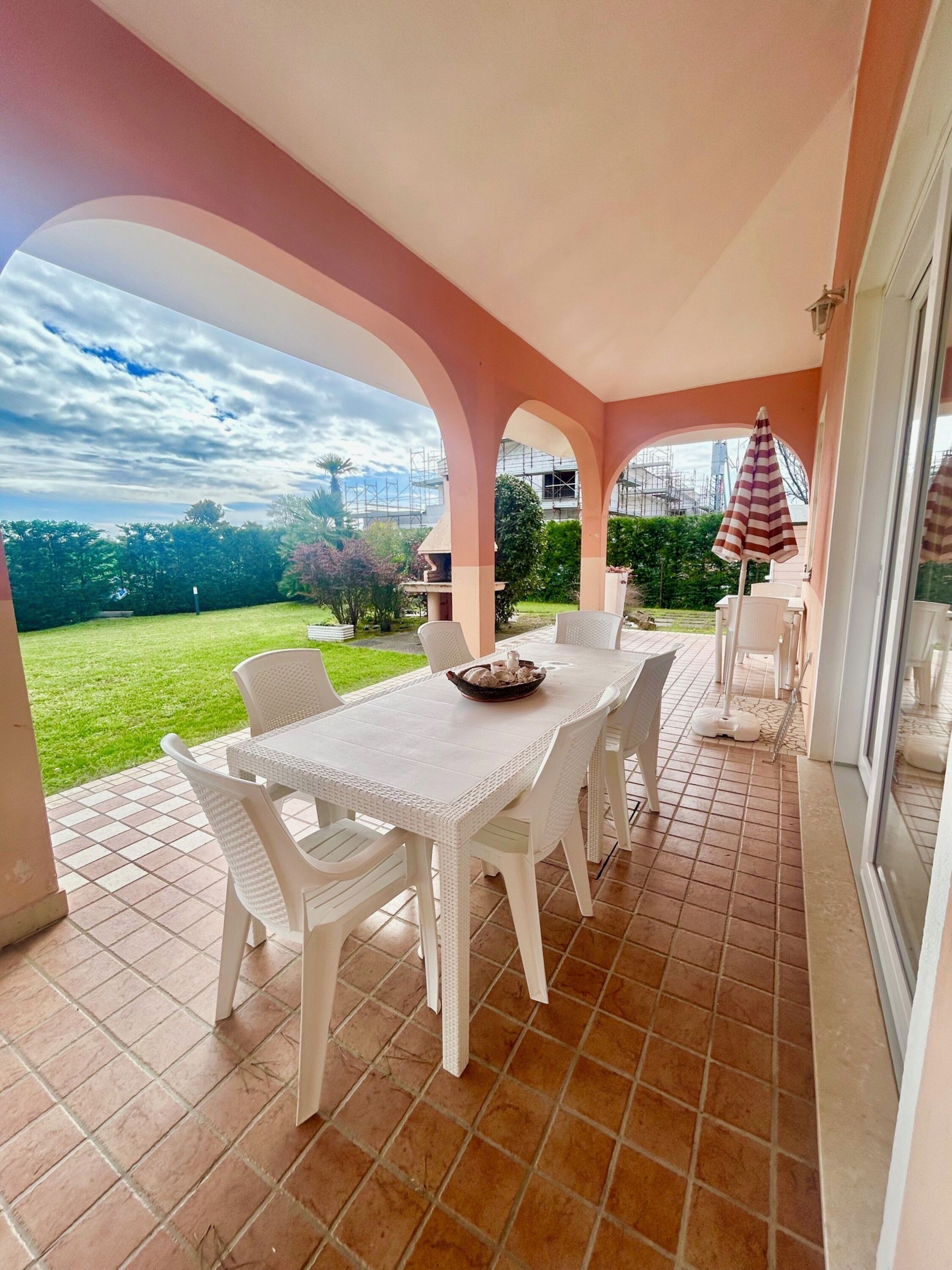 A covered patio with a white table and chairs, a striped umbrella, and a view of the surrounding greenery and buildings.