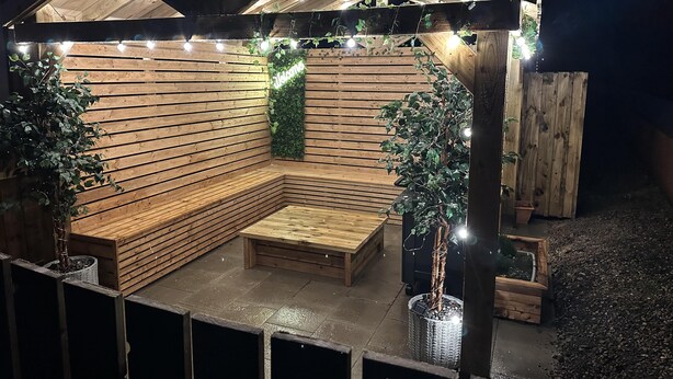 Eine Holzterrasse mit integriertem Sitzplatz und Lichterketten, umgeben von Blumentöpfen.