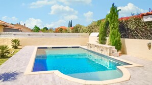 Una piscina rectangular amb un solàrium de rajoles de color clar, envoltada per una paret baixa de pedra i diferents plantes.