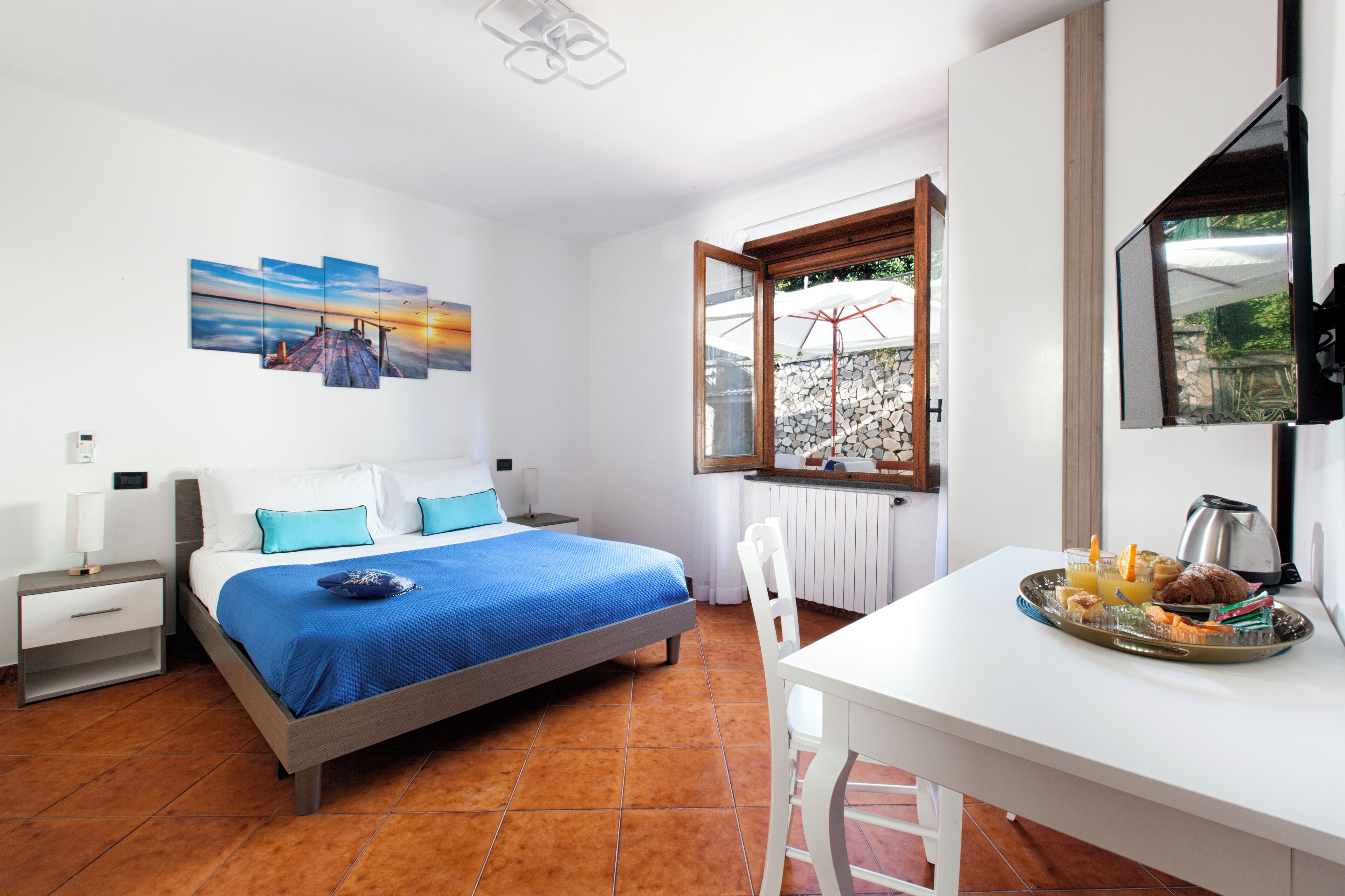 A bedroom with a bed, a nightstand, a desk with breakfast, a window, and a painting on the wall.
