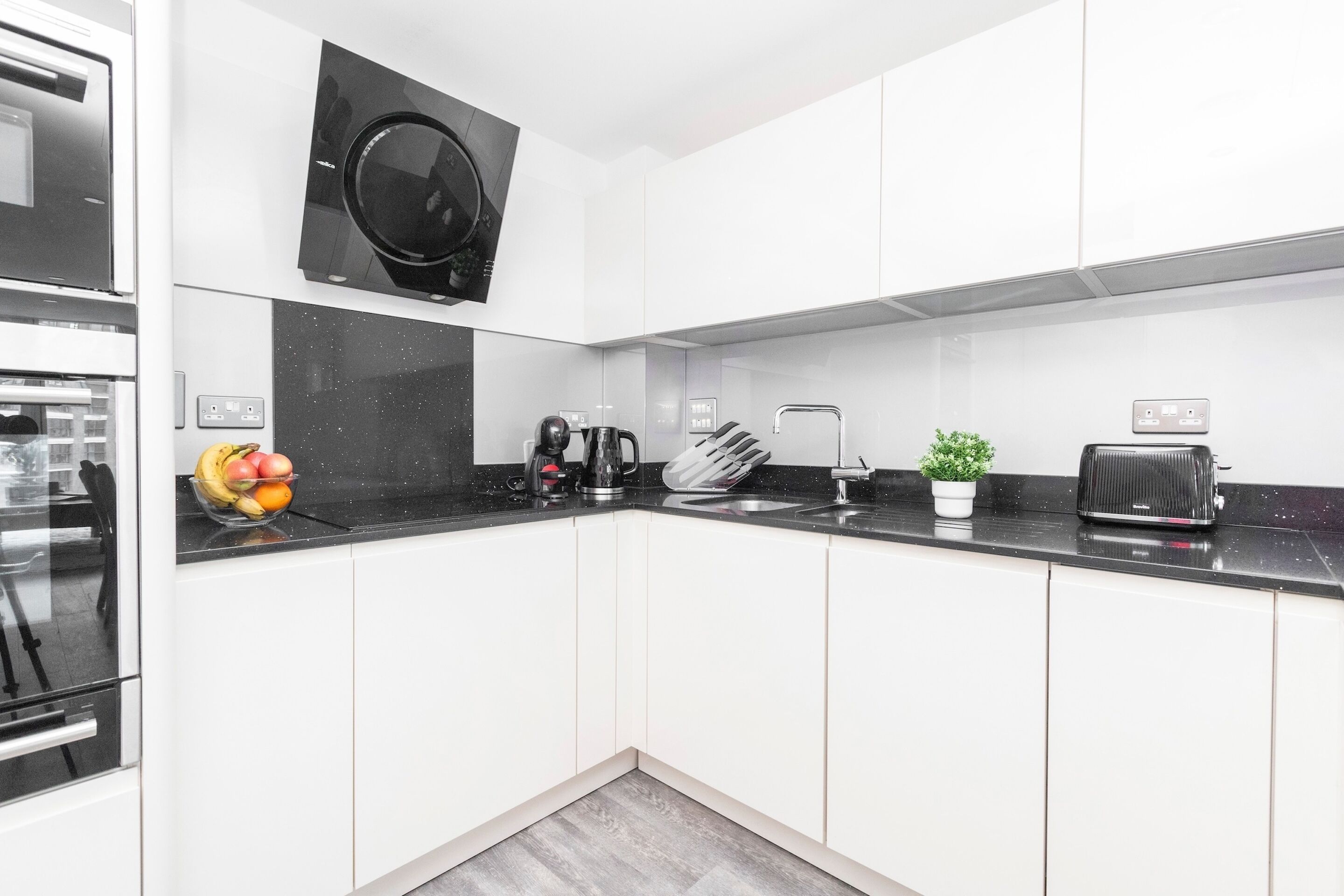 Eine moderne Küche mit weißen Schränken, einer schwarzen Arbeitsplatte, einem eingebauten Backofen, einer Kaffeemaschine, einem Toaster und einem kleinen Topfpflanzen.