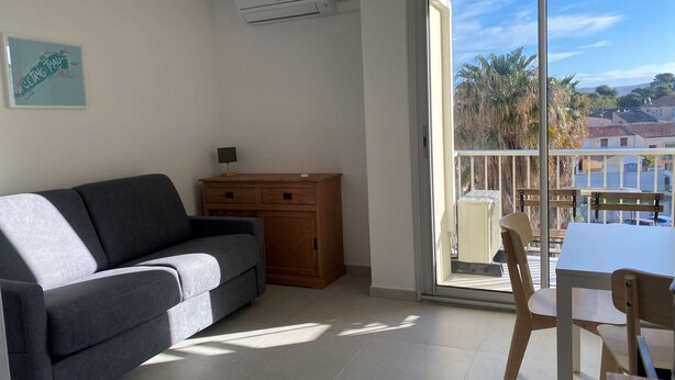 A modern living room with a grey sofa, a wooden cabinet, and a dining table with chairs. There is a large window with a view of buildings and palm trees. - Renovated Studio with Balcony, A/C, Wi-Fi and Modern Amenities - 5 Min Walk to Thermes (Balaruc-les-Bains)