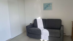 A grey sofa in a room with white walls and a tiled floor. - Renovated Studio with Balcony, A/C, Wi-Fi and Modern Amenities - 5 Min Walk to Thermes (Balaruc-les-Bains)